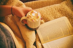 Picture of a person curled up under a blanket with hot chocolate and a book.